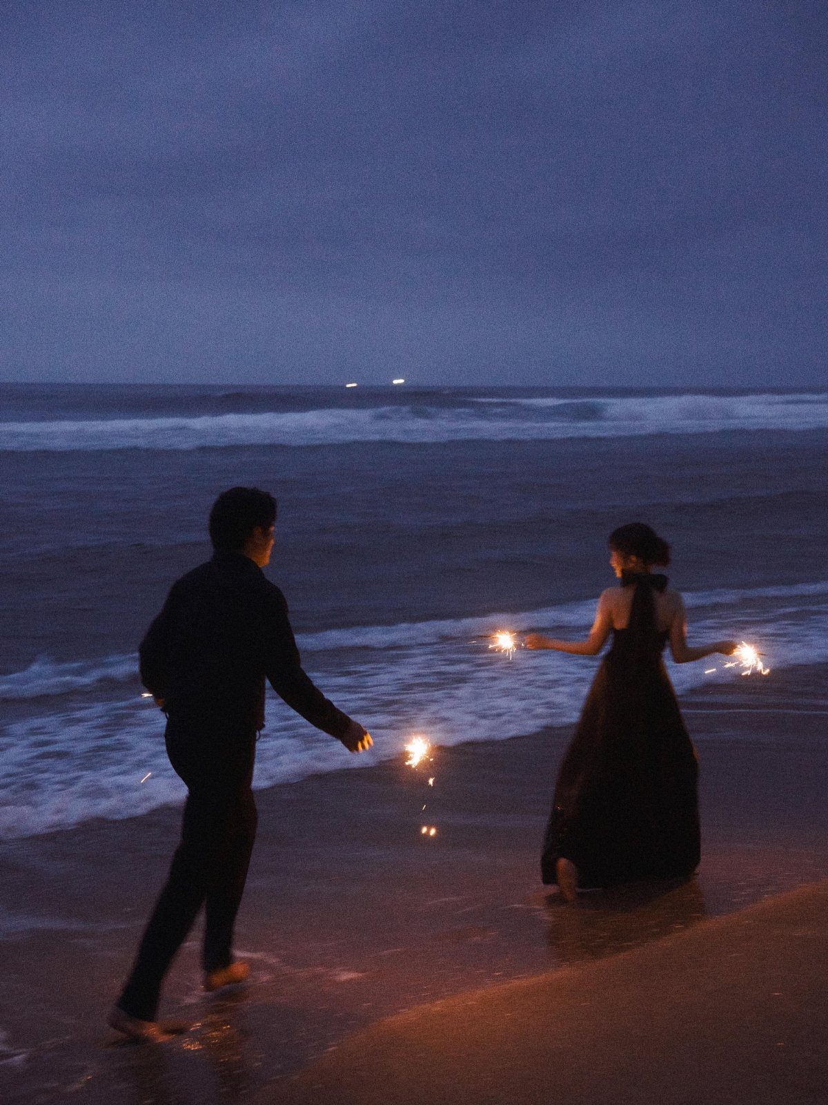 Couple holding lit sparklers on a beach at dusk by the ocean waves