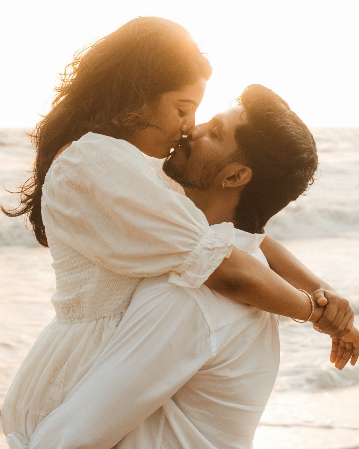Man carrying woman in white clothing on a beach during a sunset photoshoot