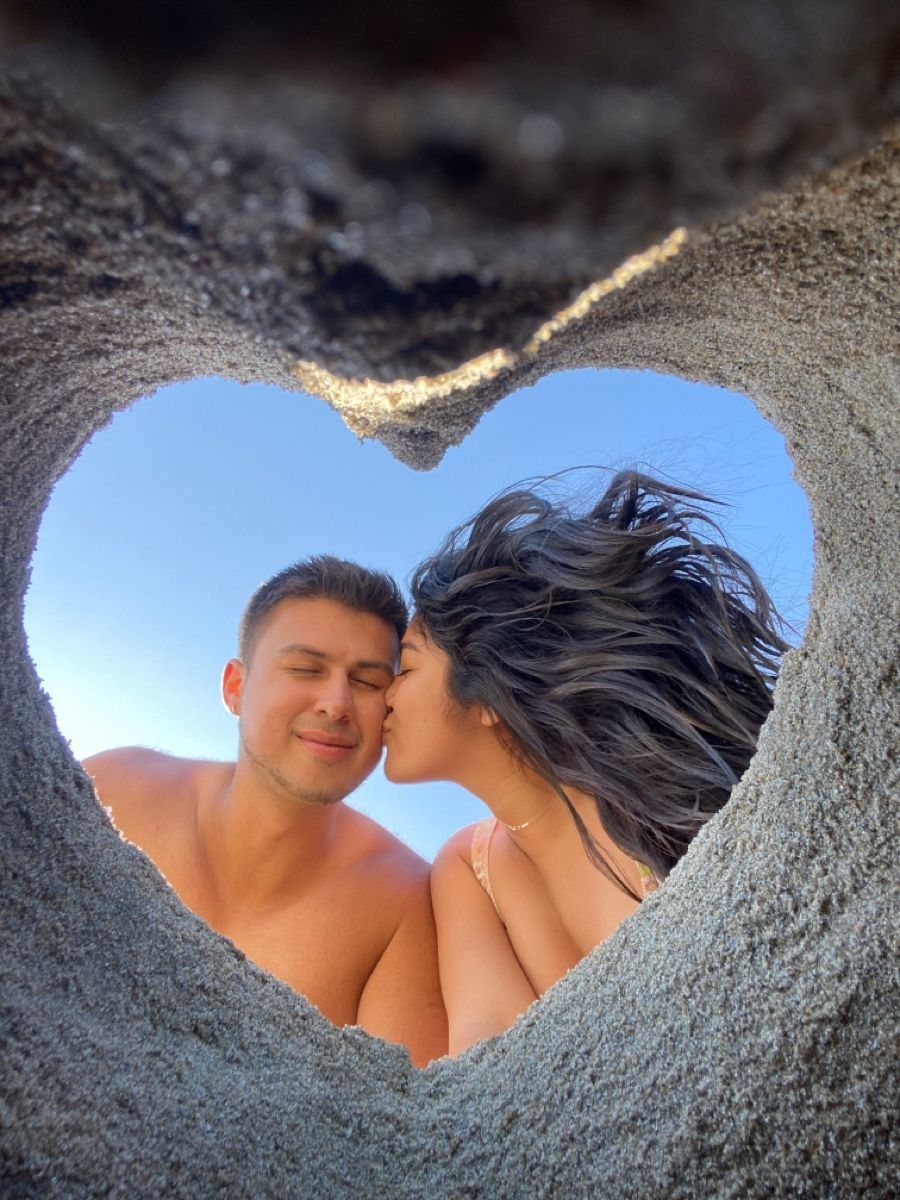 Couple kissing viewed through a heart-shaped hole carved in beach sand.