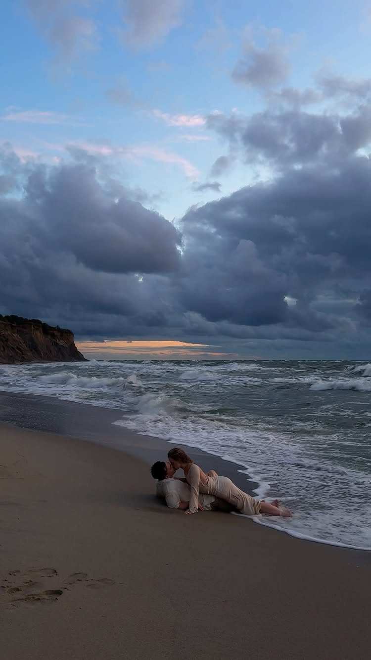 Couple lying on a sandy beach with waves washing over them under cloudy skies.