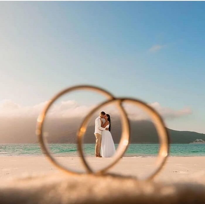Two gold wedding rings framing a couple embracing on a sunny white beach.