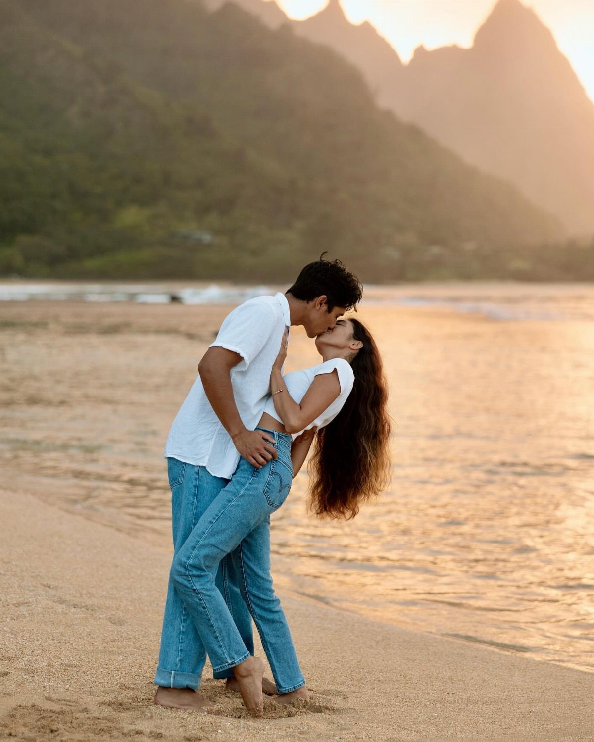 Couple in white shirts and jeans kissing while dipping on a sandy beach.