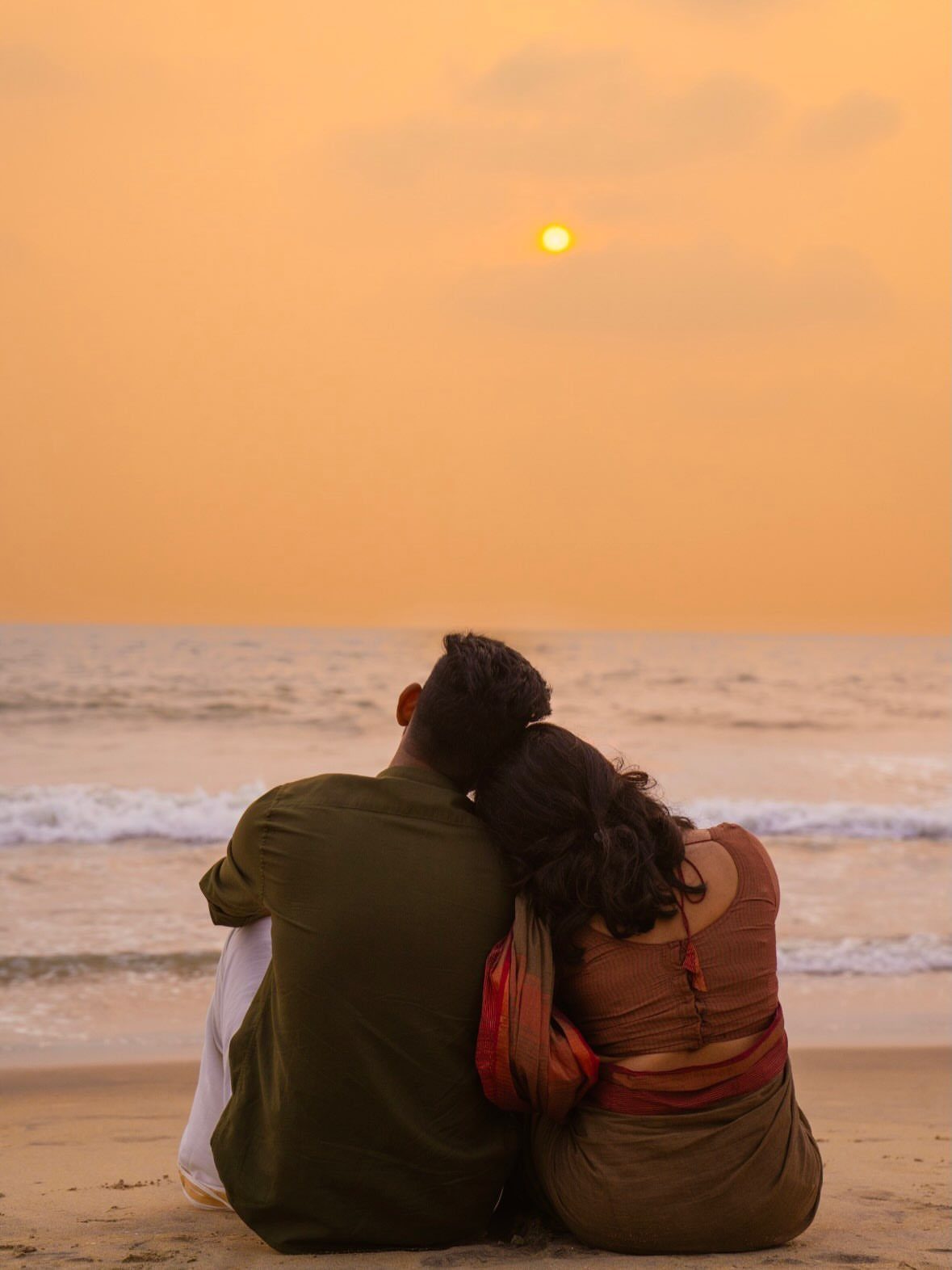 Couple sitting on the sand watching a golden sunset over the ocean waves.