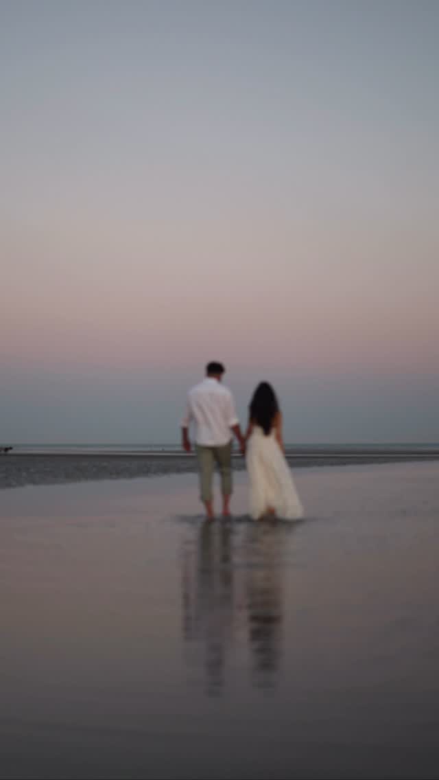 Couple walking hand-in-hand on a wet beach during a soft purple sunset.