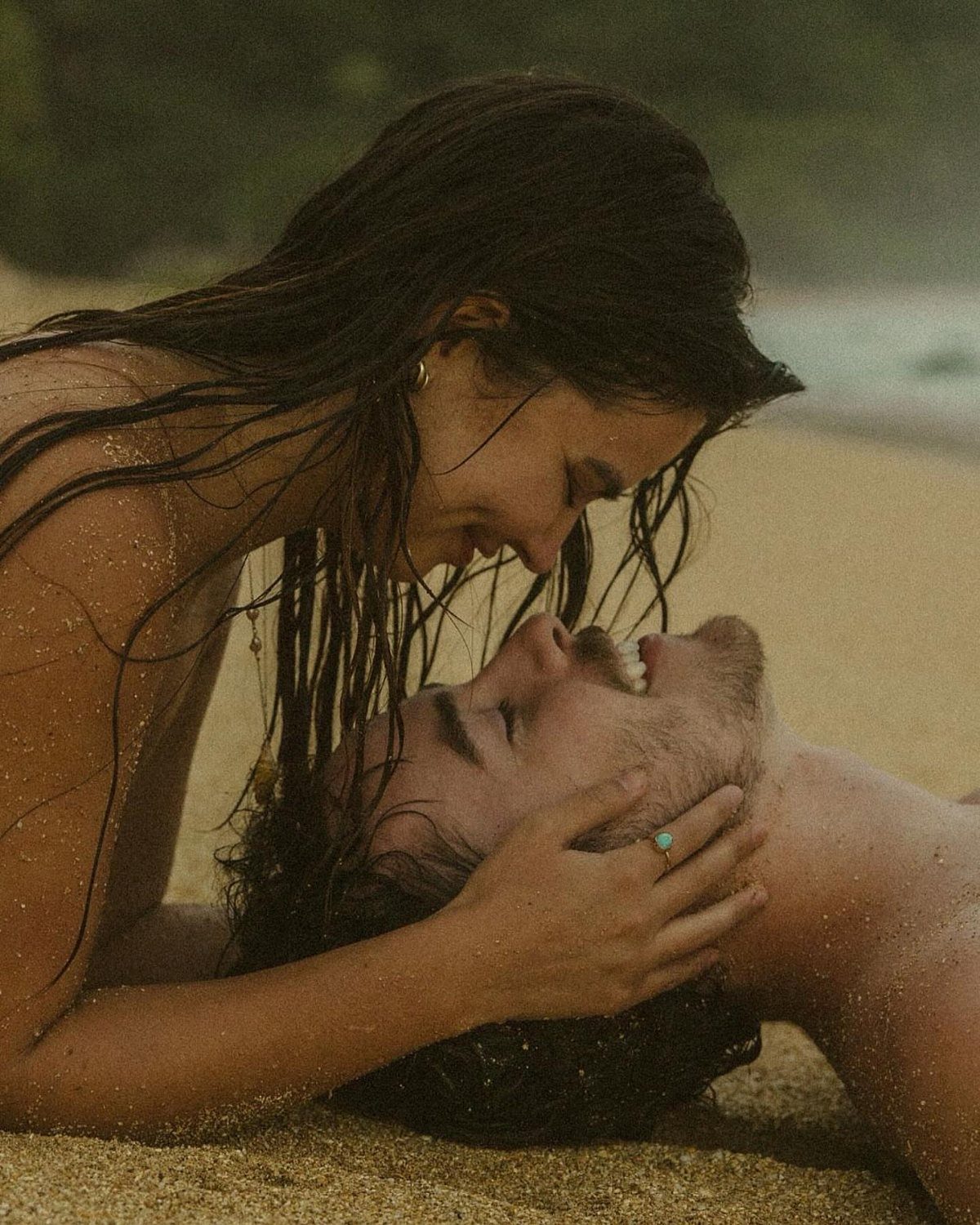 Couple with wet hair lying on sandy beach, woman cradling man's face.