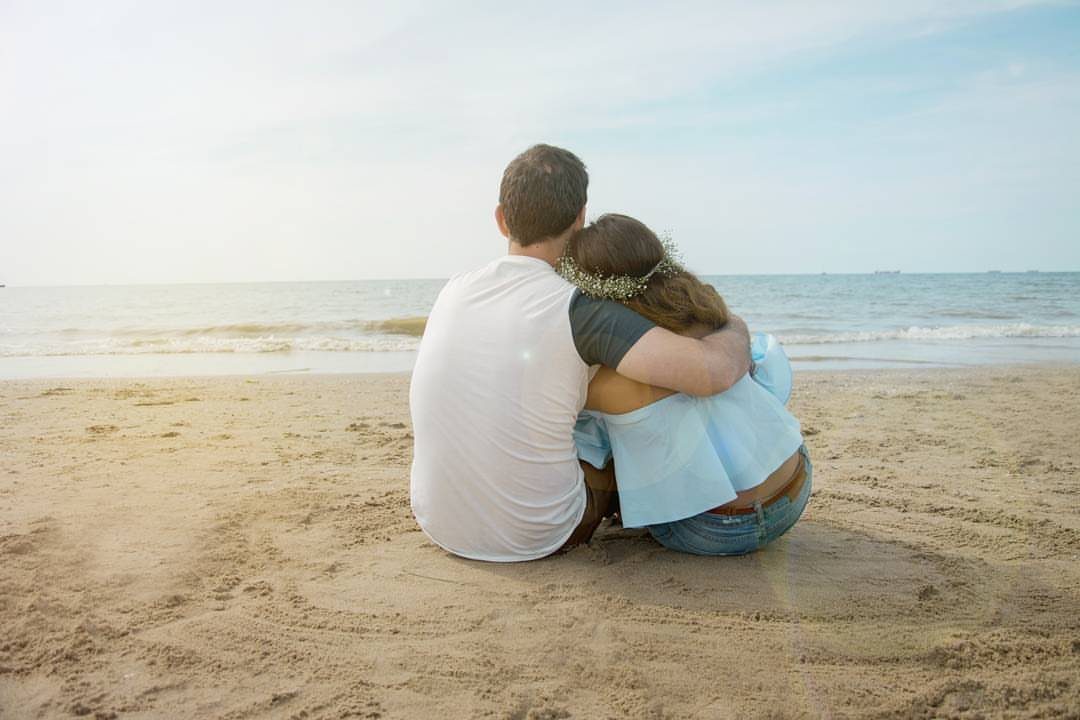 Couple sitting on a sandy beach hugging while looking out at the ocean