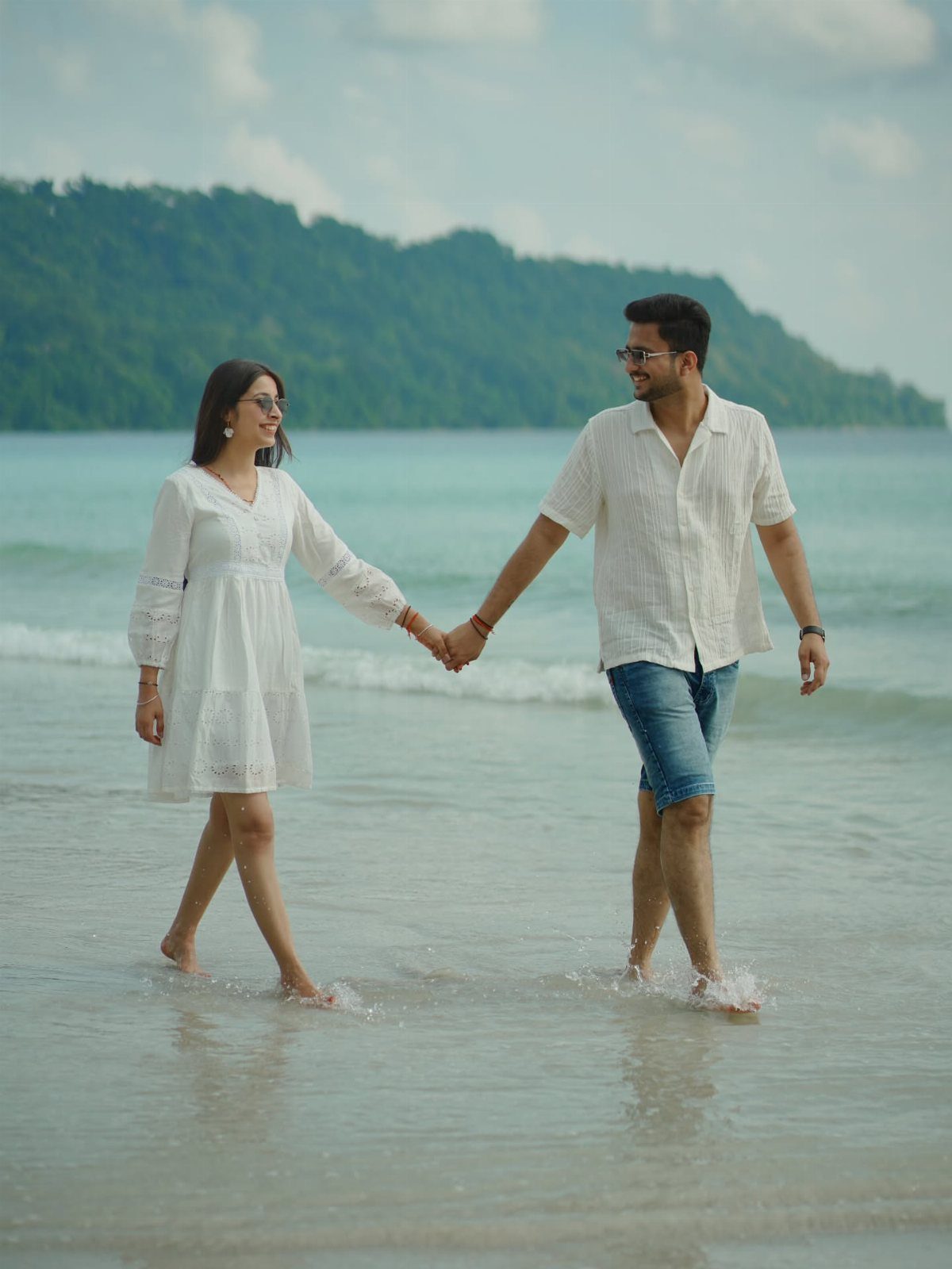 Couple in white clothing walking hand-in-hand through shallow ocean water on a beach.