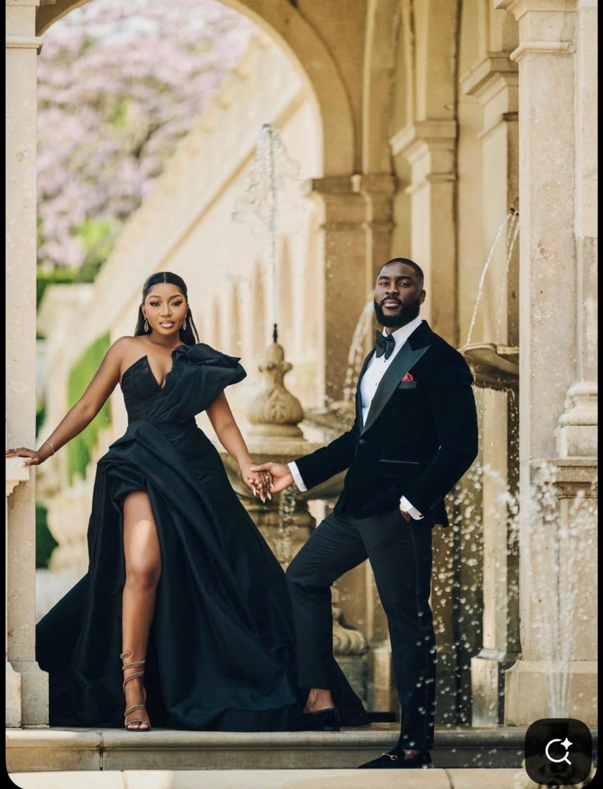 Black couple posing in a black velvet tuxedo and one-shoulder gown by a fountain.