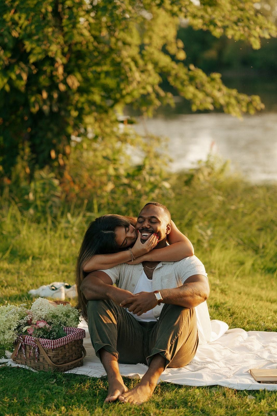 Black couple hugging on a white blanket in grass by a river with picnic basket
