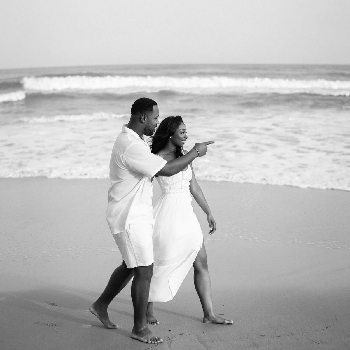 Black couple in white outfits walking on a sandy beach by the ocean.