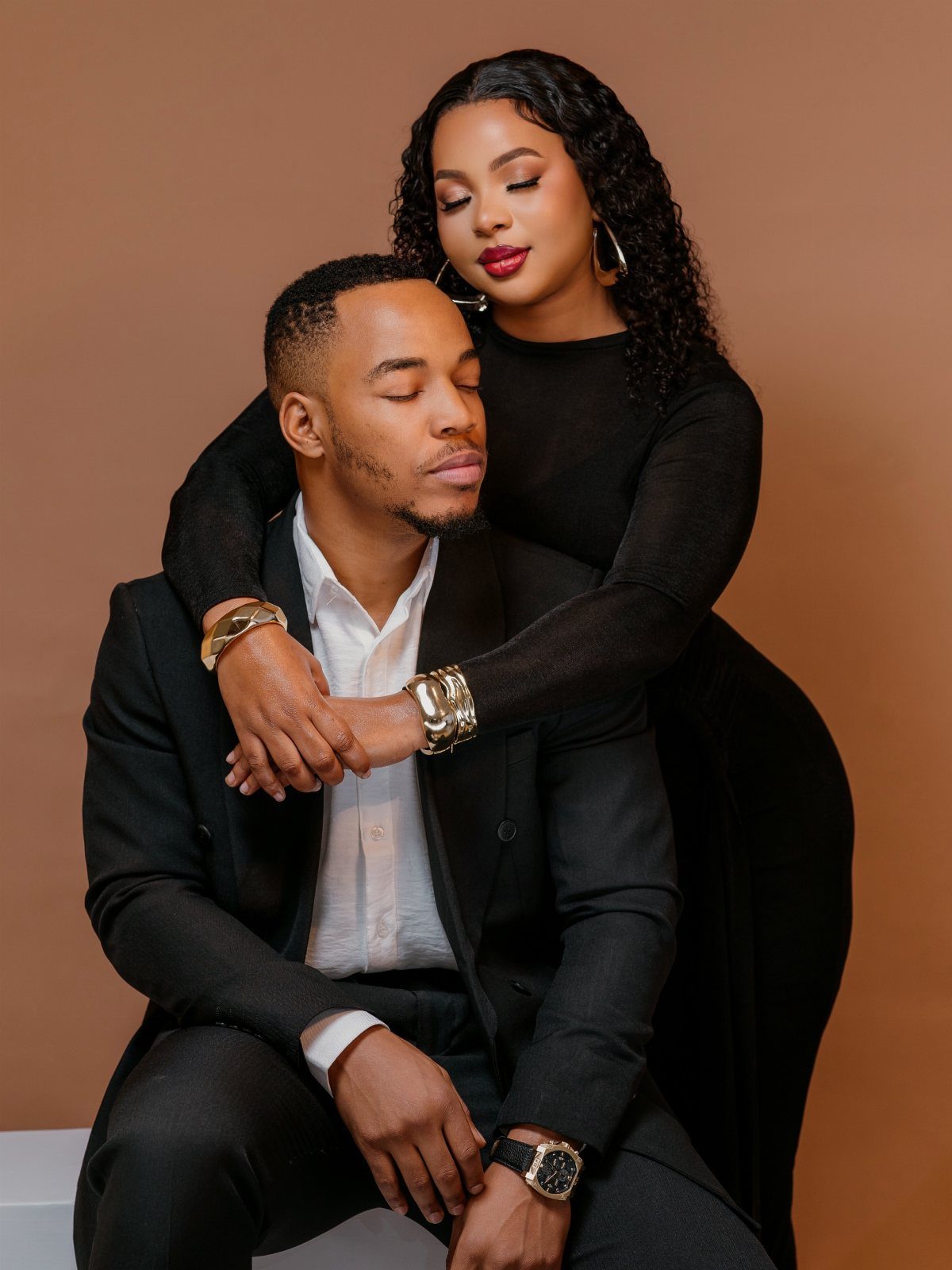Black couple in formal black attire posing romantically against a tan background.