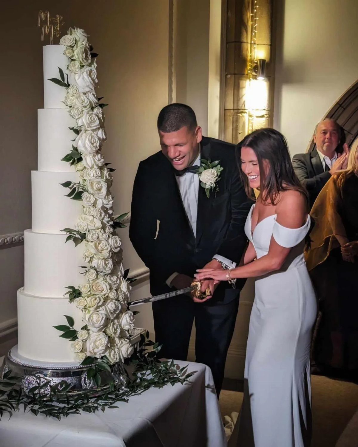 Bride and groom smiling while cutting a five-tier white rose wedding cake together.