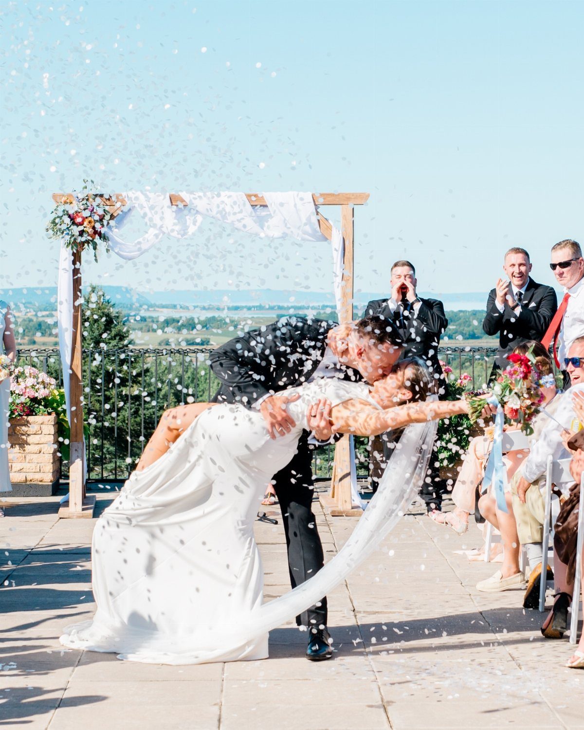 Bride and groom kissing in a dip pose while white confetti falls around them.