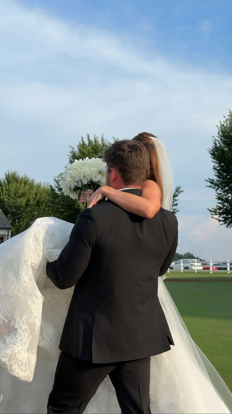 Groom in black suit carrying bride in white lace wedding dress outdoors