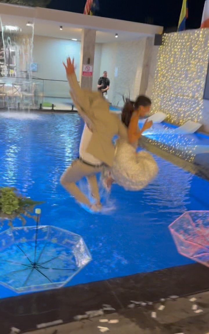 Groom in tan suit jumping into a pool while carrying a bride.
