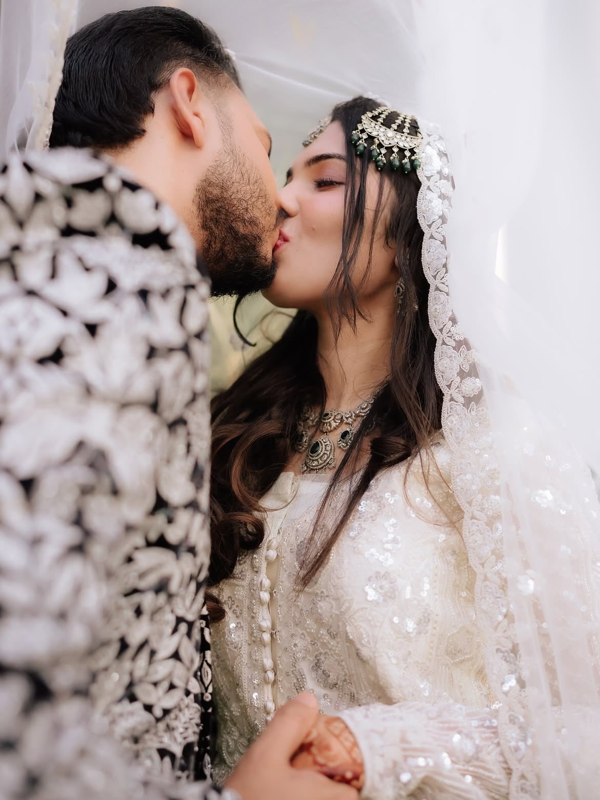 Bride and groom in traditional white wedding attire kissing under a sheer veil.