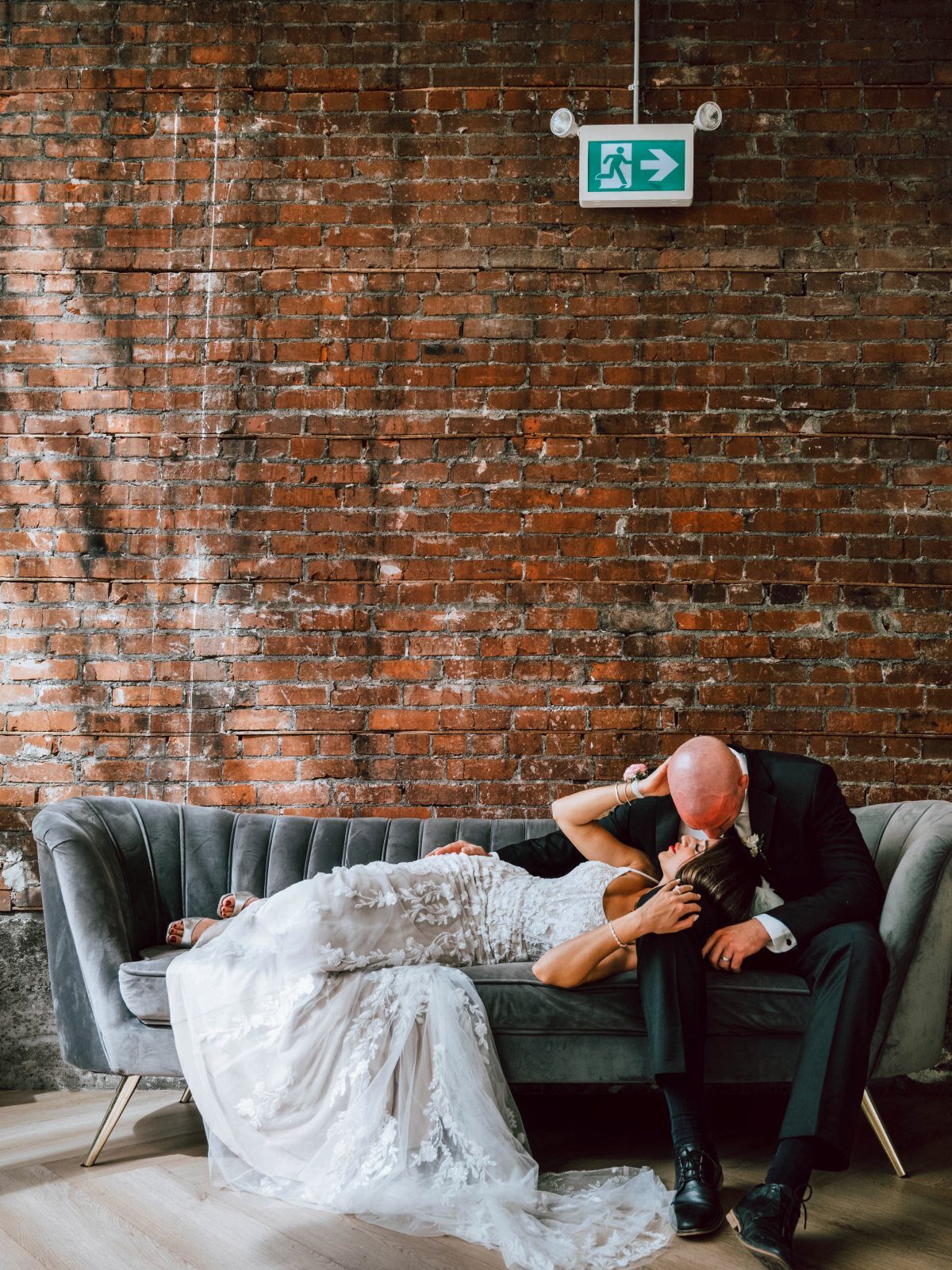 Bride and groom kissing on a grey velvet sofa against a brick wall