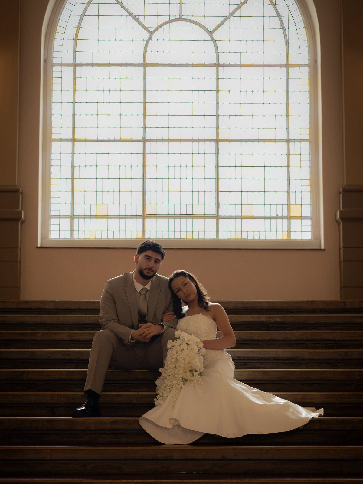 Bride and groom sitting on wooden stairs with a large arched window behind them