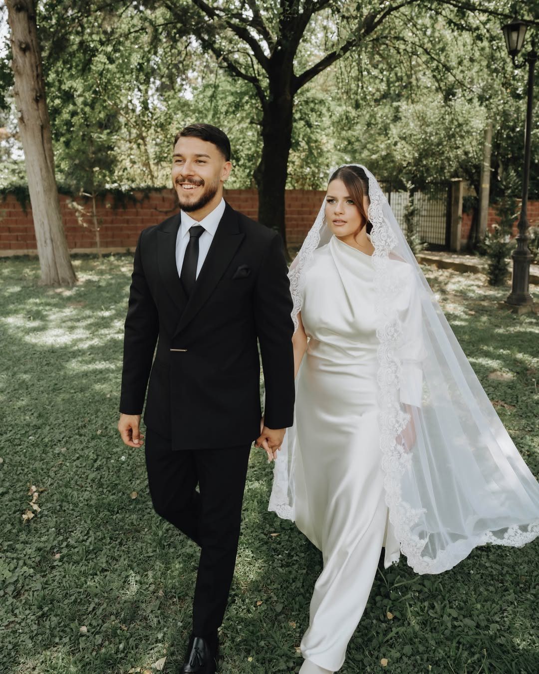 Bride and groom walking hand in hand across a sunny green lawn.