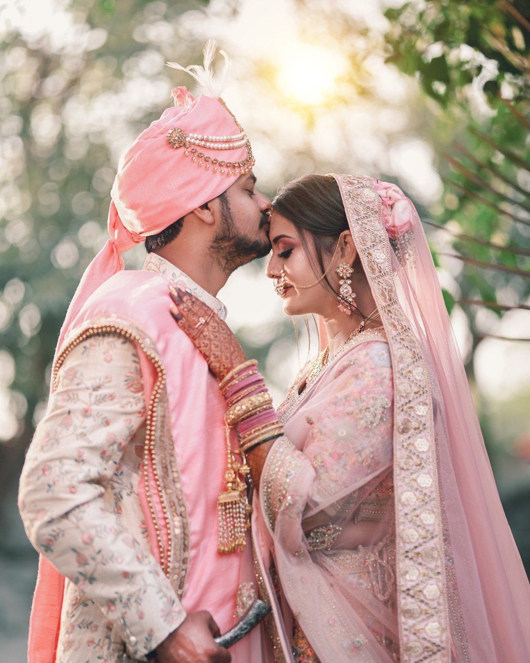 Groom in pink turban kissing the bride's forehead in traditional Indian wedding attire.