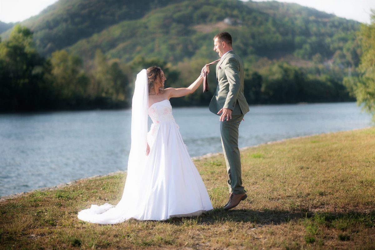 Bride in white gown playfully pulling the groom's tie by a riverbank.