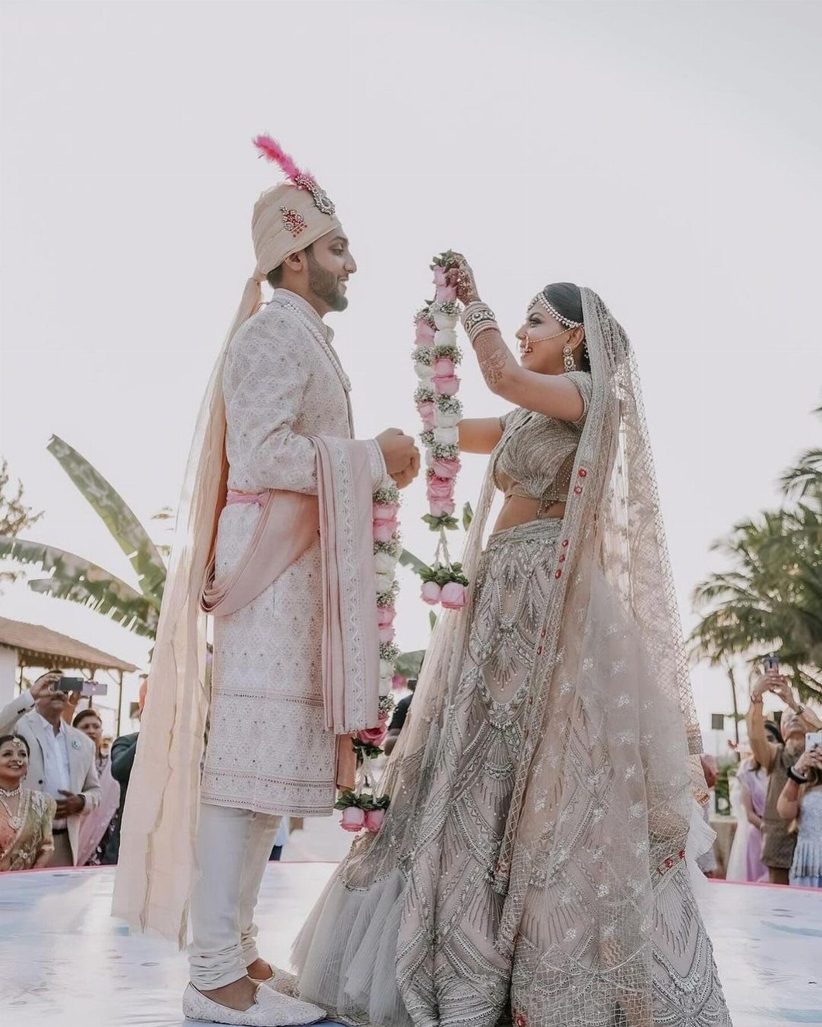 Bride and groom in traditional Indian wedding attire exchanging floral garlands outdoors.