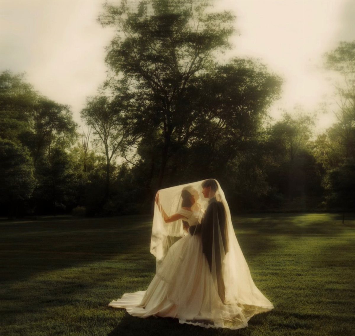 Bride and groom standing close together under a white veil in a park