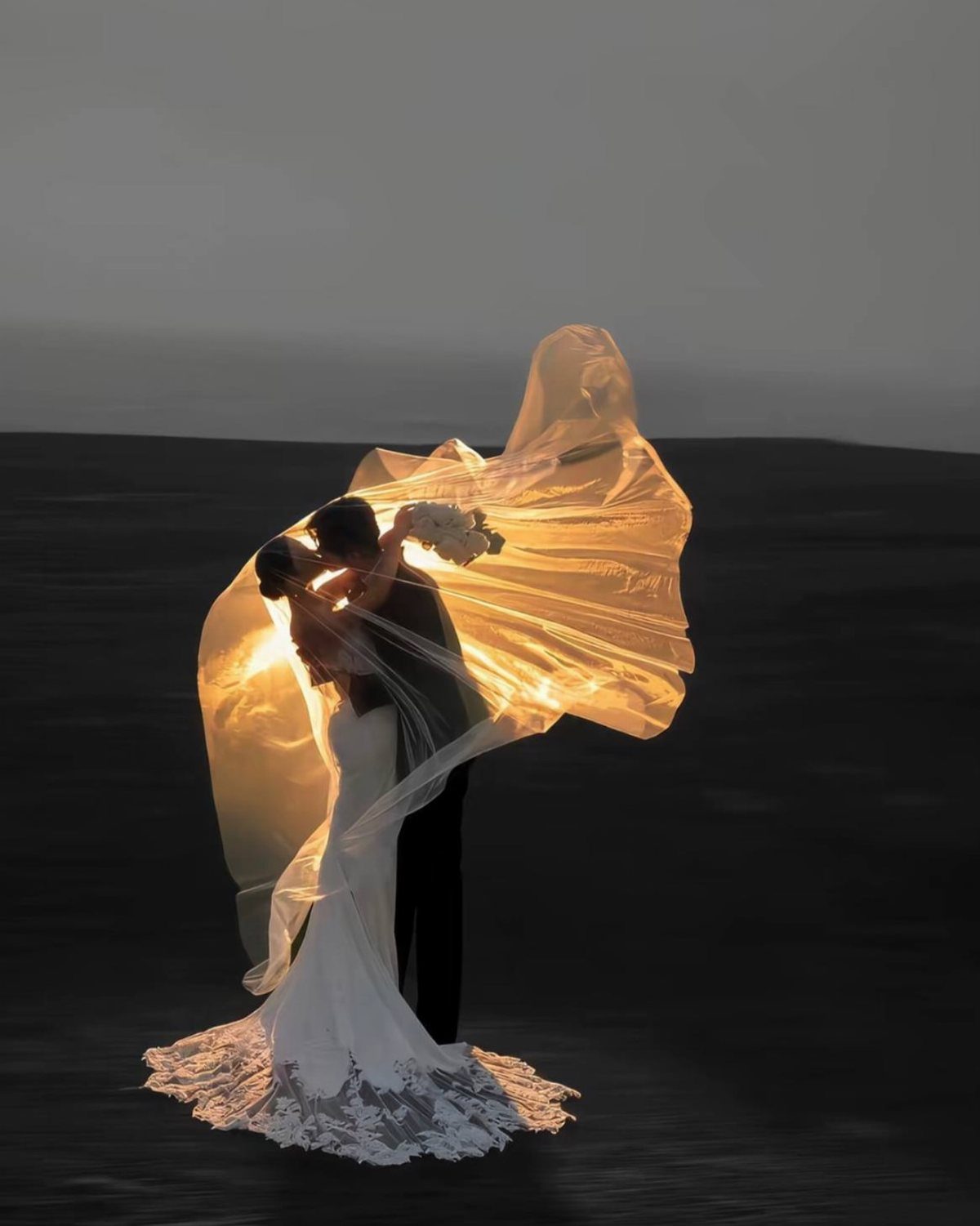 Bride and groom kissing under a flowing veil during a golden sunset.