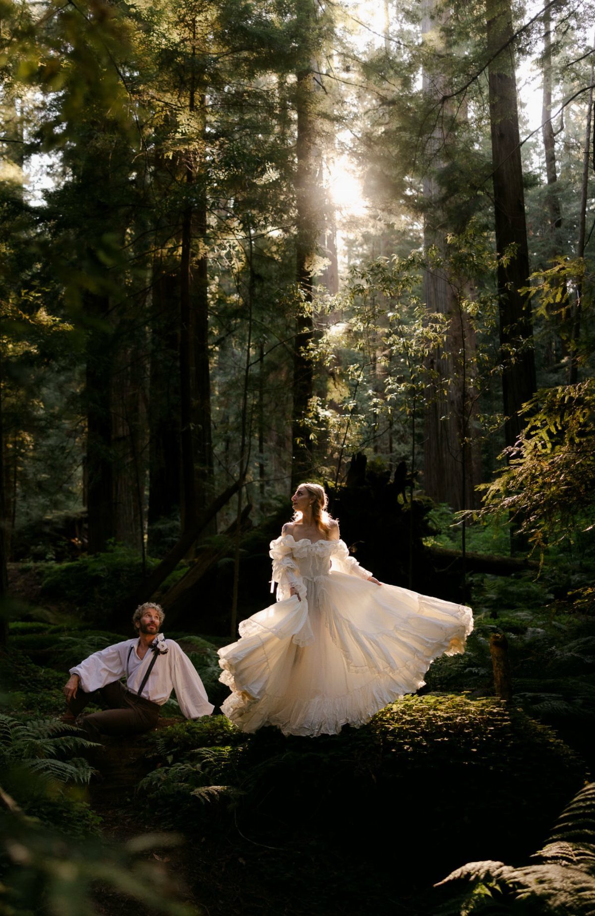 Bride in white gown and groom posing among tall trees in a sunlit forest