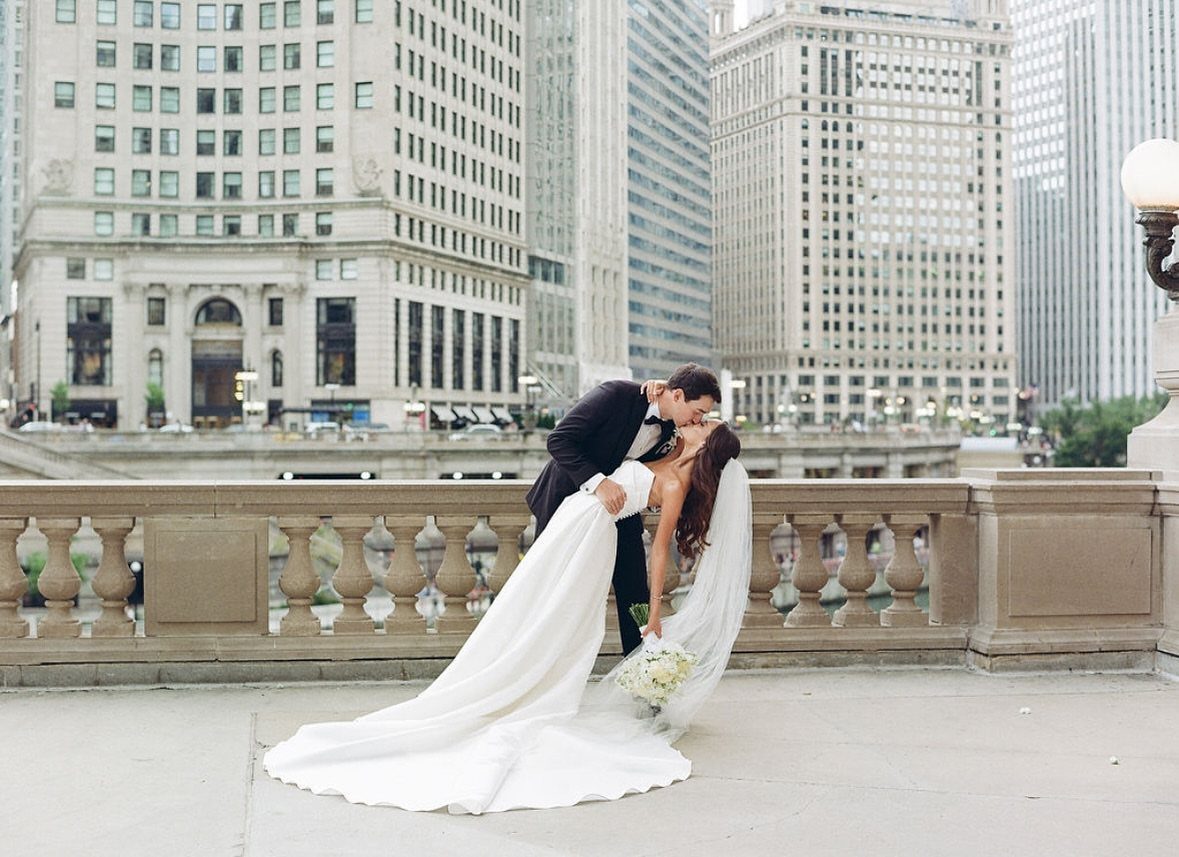 Groom dipping bride in white gown for a kiss on a city bridge