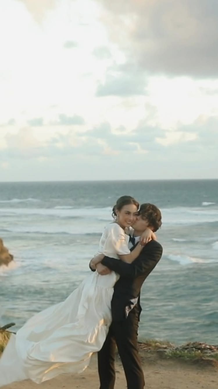 Bride and groom embracing on a cliff overlooking a vast, crashing ocean.