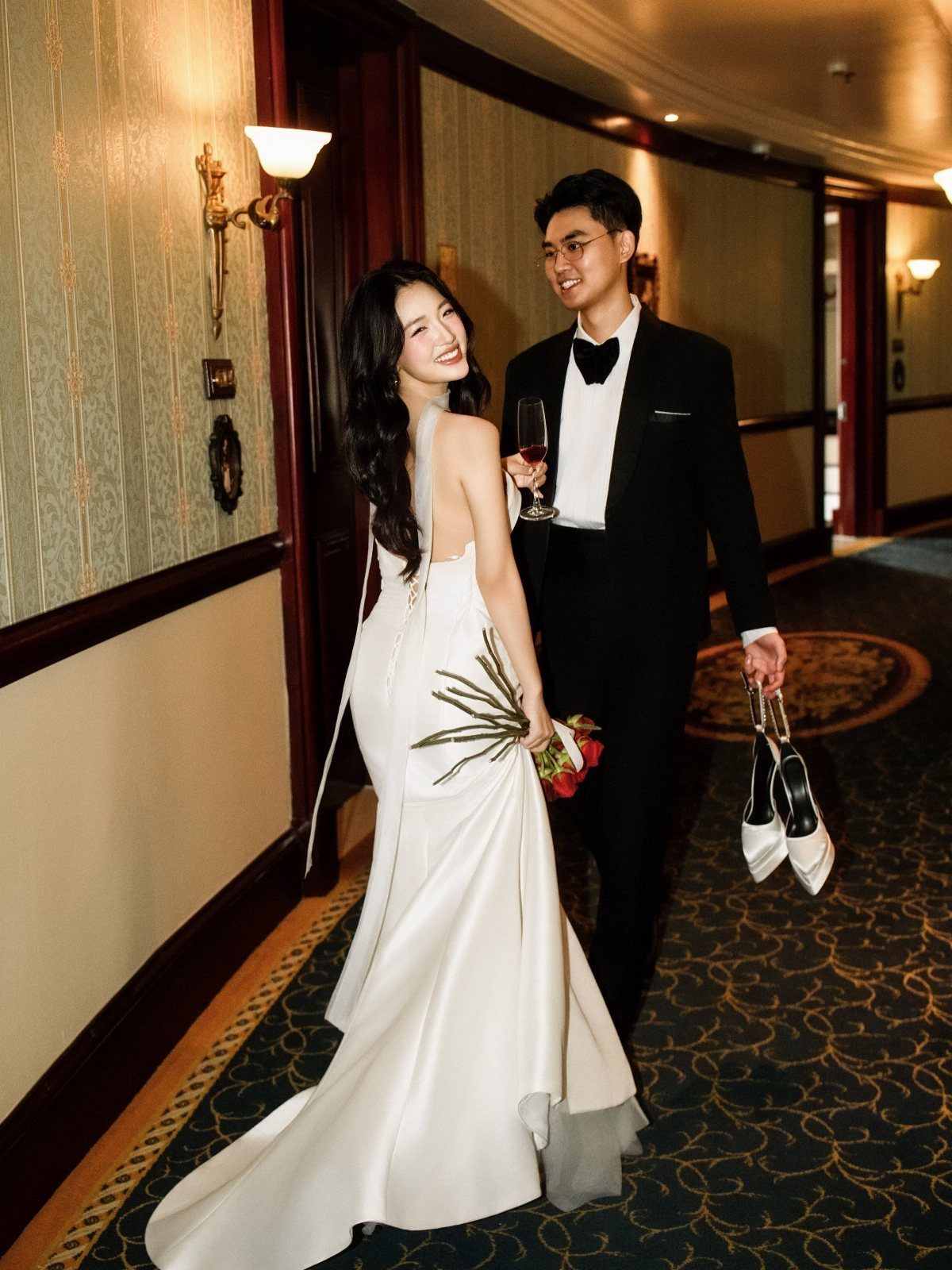Bride and groom walking in a hotel hallway, groom carrying bride's white shoes.