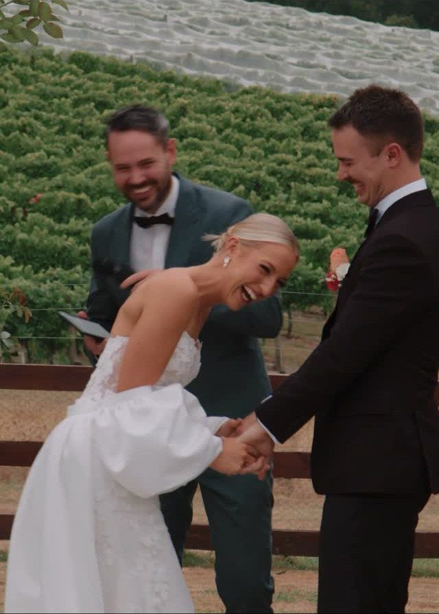 Bride and groom laughing while holding hands with an officiant in a vineyard