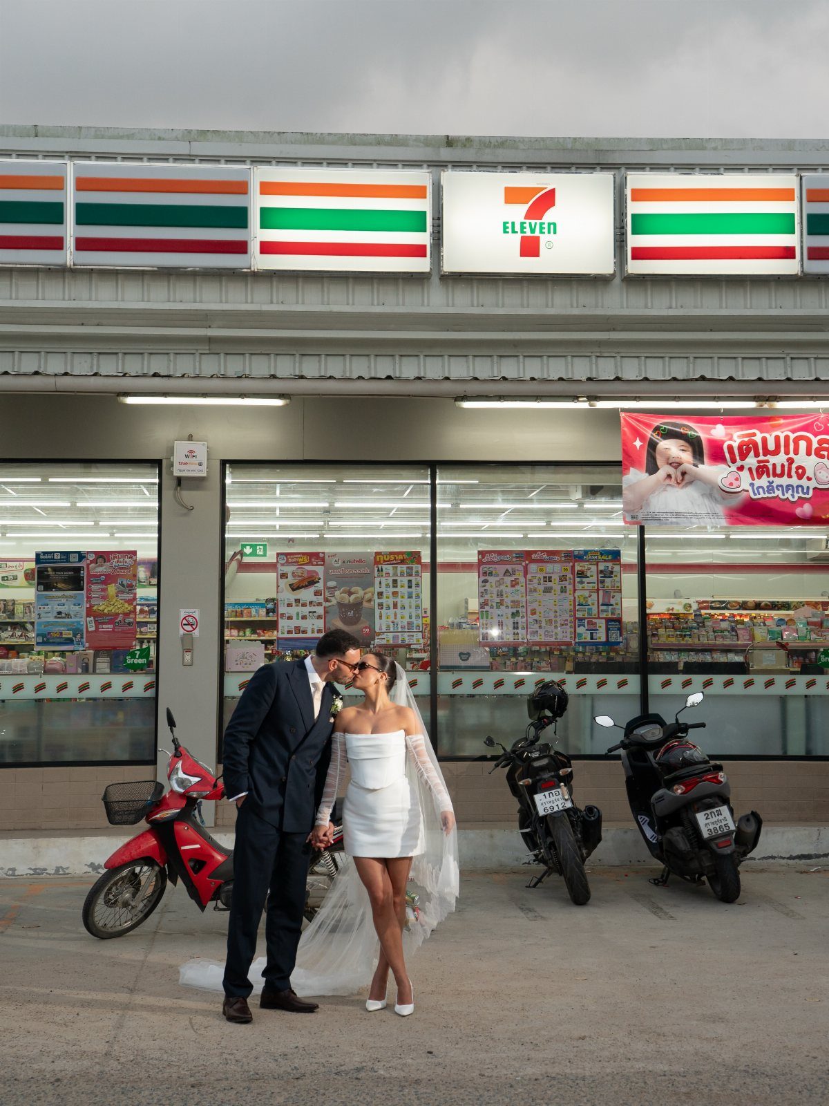 Bride and groom kissing in front of a 7-Eleven convenience store with motorcycles.
