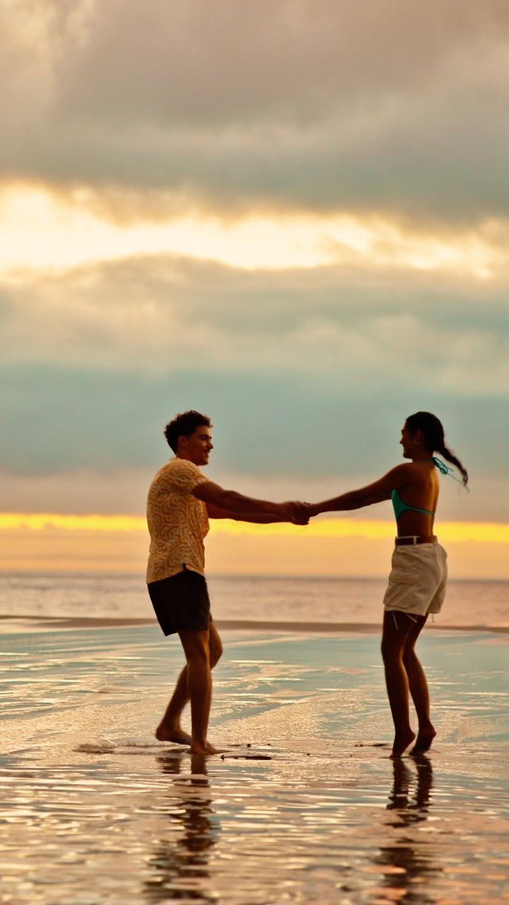Couple holding hands and dancing in shallow beach water during a golden sunset.