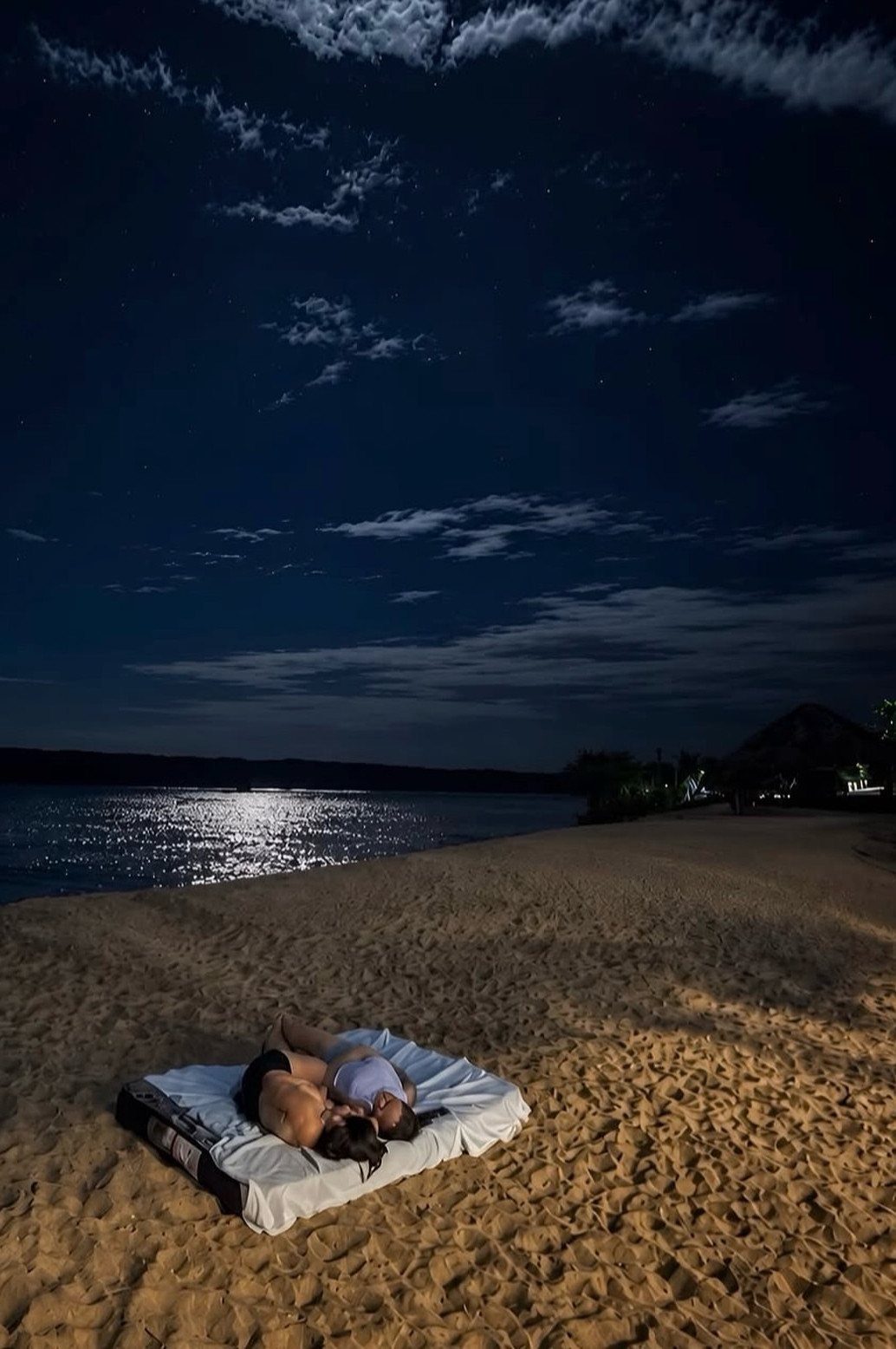 Couple lying on a white blanket on a sandy beach under a starry night sky