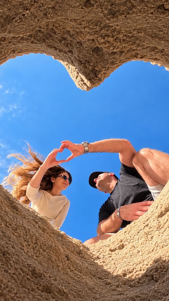 Couple posing inside a heart-shaped sand frame making a heart with their hands.