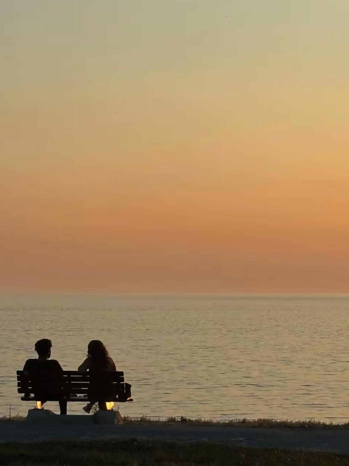 Silhouetted couple sitting on a bench overlooking the ocean during a golden sunset.