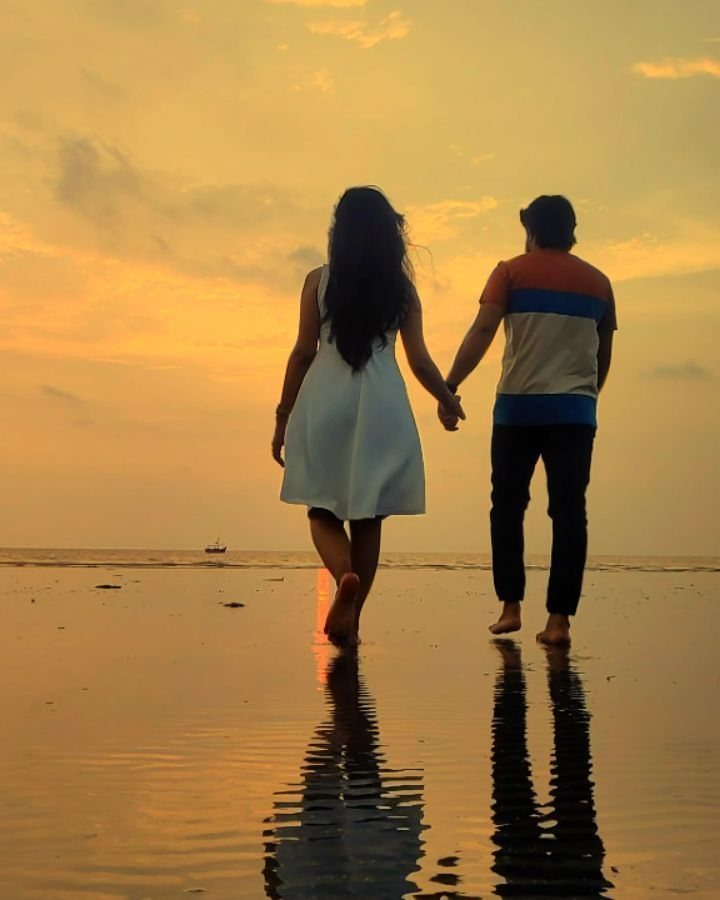 Couple walking hand-in-hand on a beach during a golden sunset with reflections.