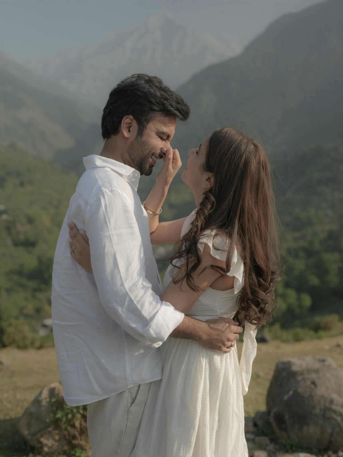 Couple in white outfits embracing in front of a mountainous landscape.