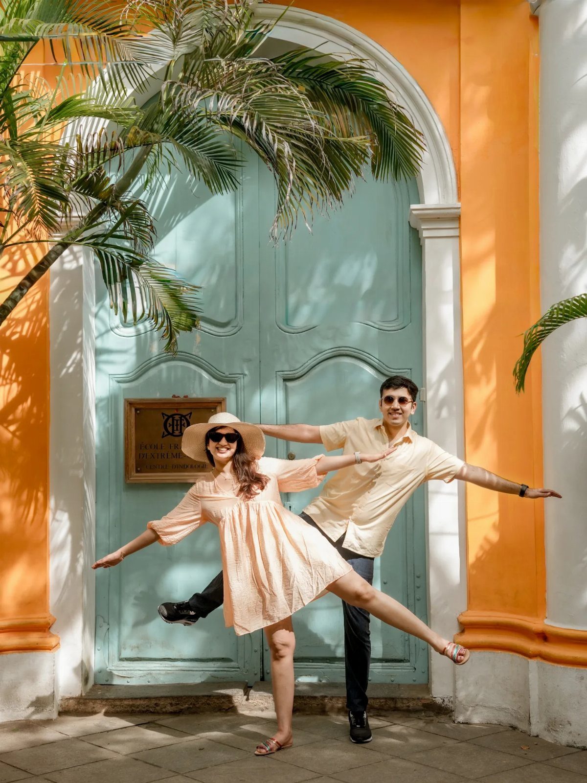 Couple in light summer outfits posing before a blue door and orange wall.