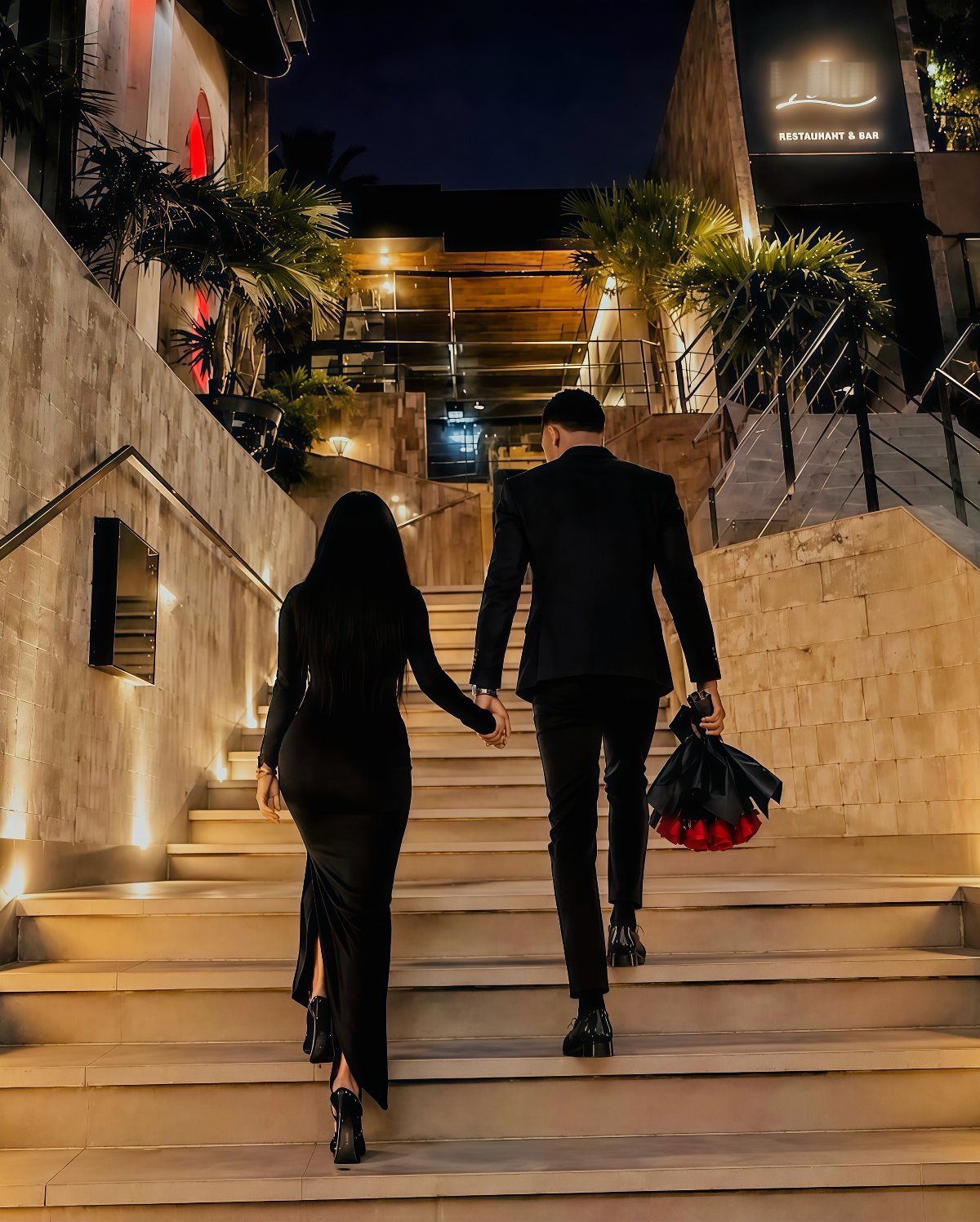 Couple in formal black attire walking up lit stairs with red roses.