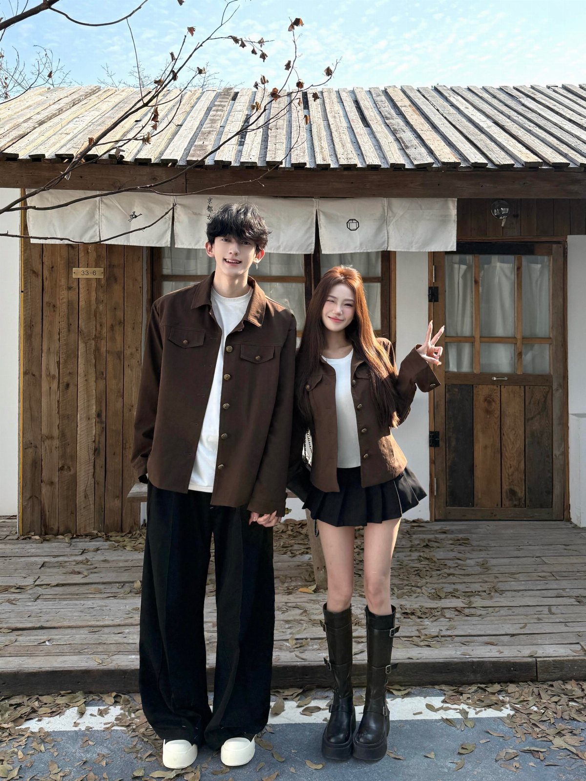 Couple posing in matching brown jackets and white tops before a wooden house.