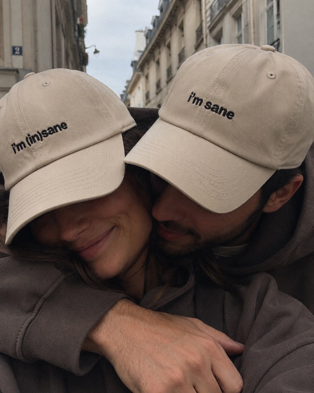 Couple wearing matching beige baseball caps and brown hoodies embracing in a city.