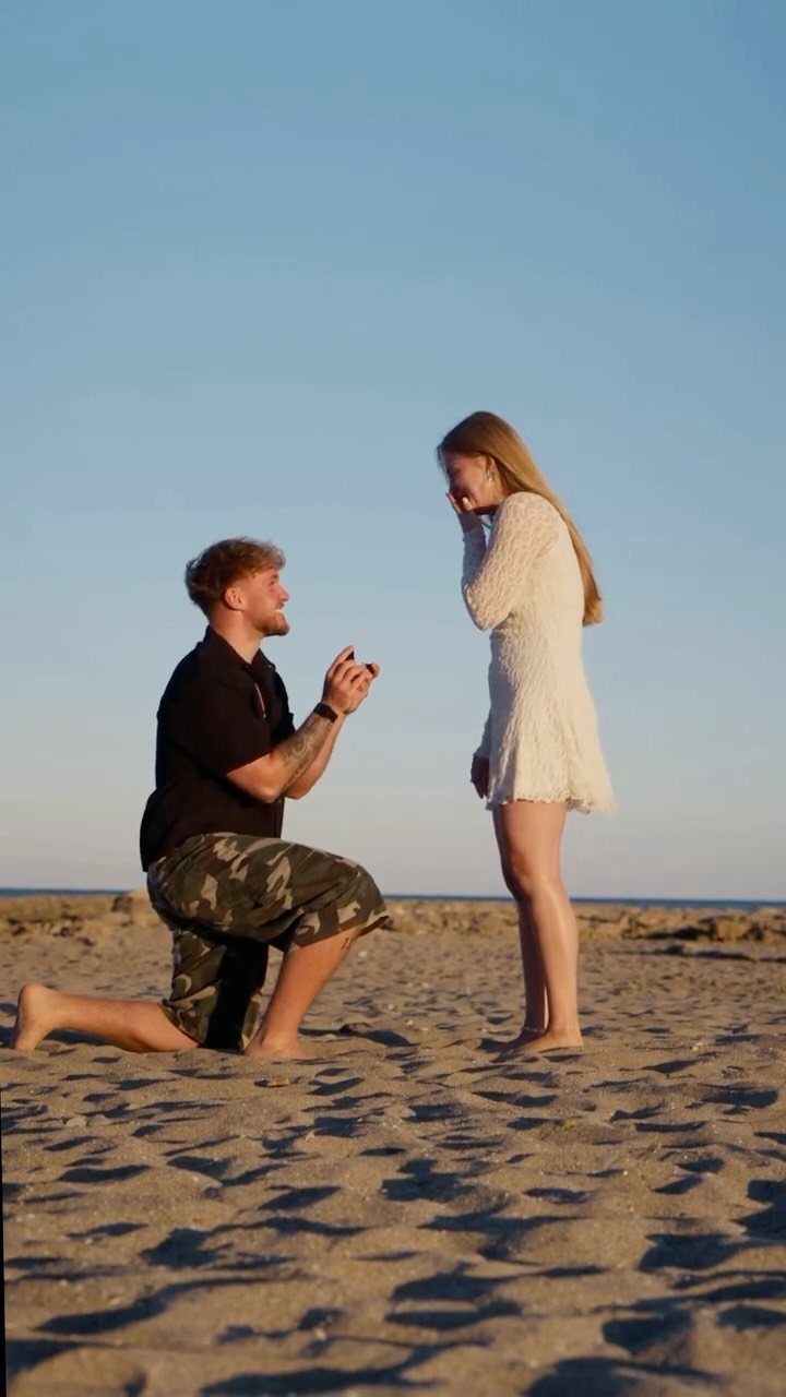 Man kneeling on a sandy beach proposing to a woman in a white dress.