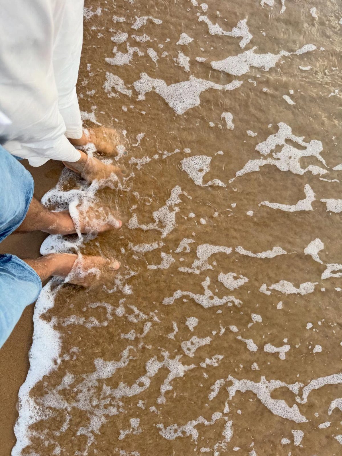 Two people's feet standing in foamy ocean water on a sandy beach.