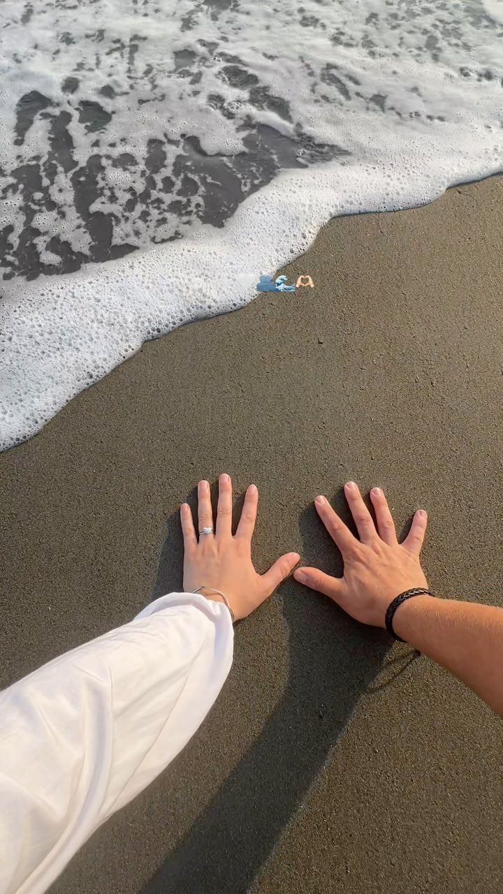 Two hands resting on dark beach sand as white sea foam washes ashore.