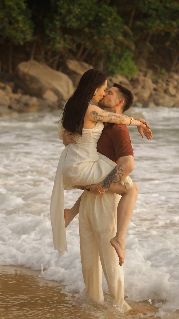 Man lifting woman in white dress while kissing in beach surf