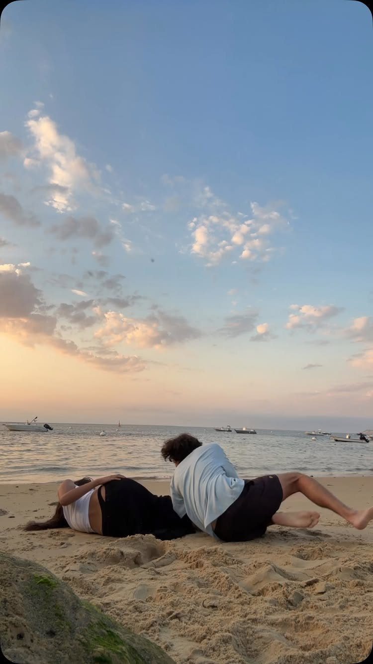 Couple lying on a sandy beach at sunset with boats in the distance.