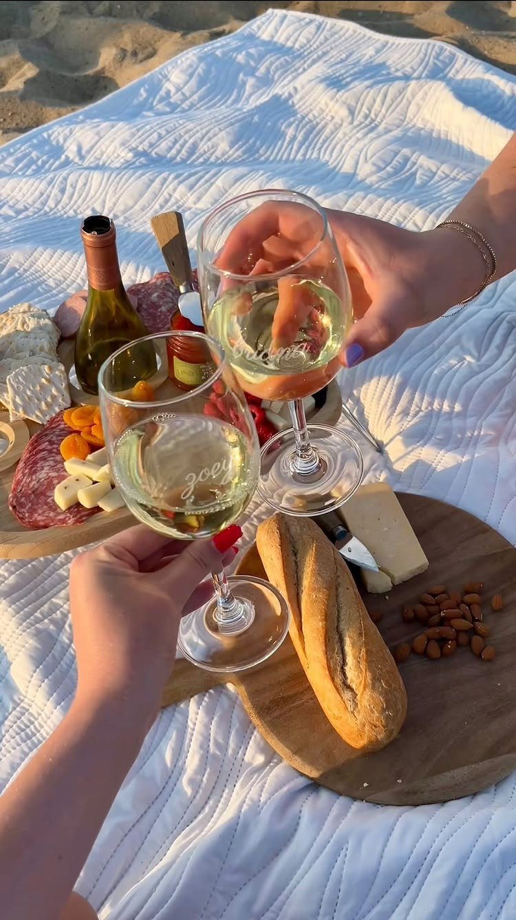 Two people clinking wine glasses over a gourmet picnic spread on a beach blanket.